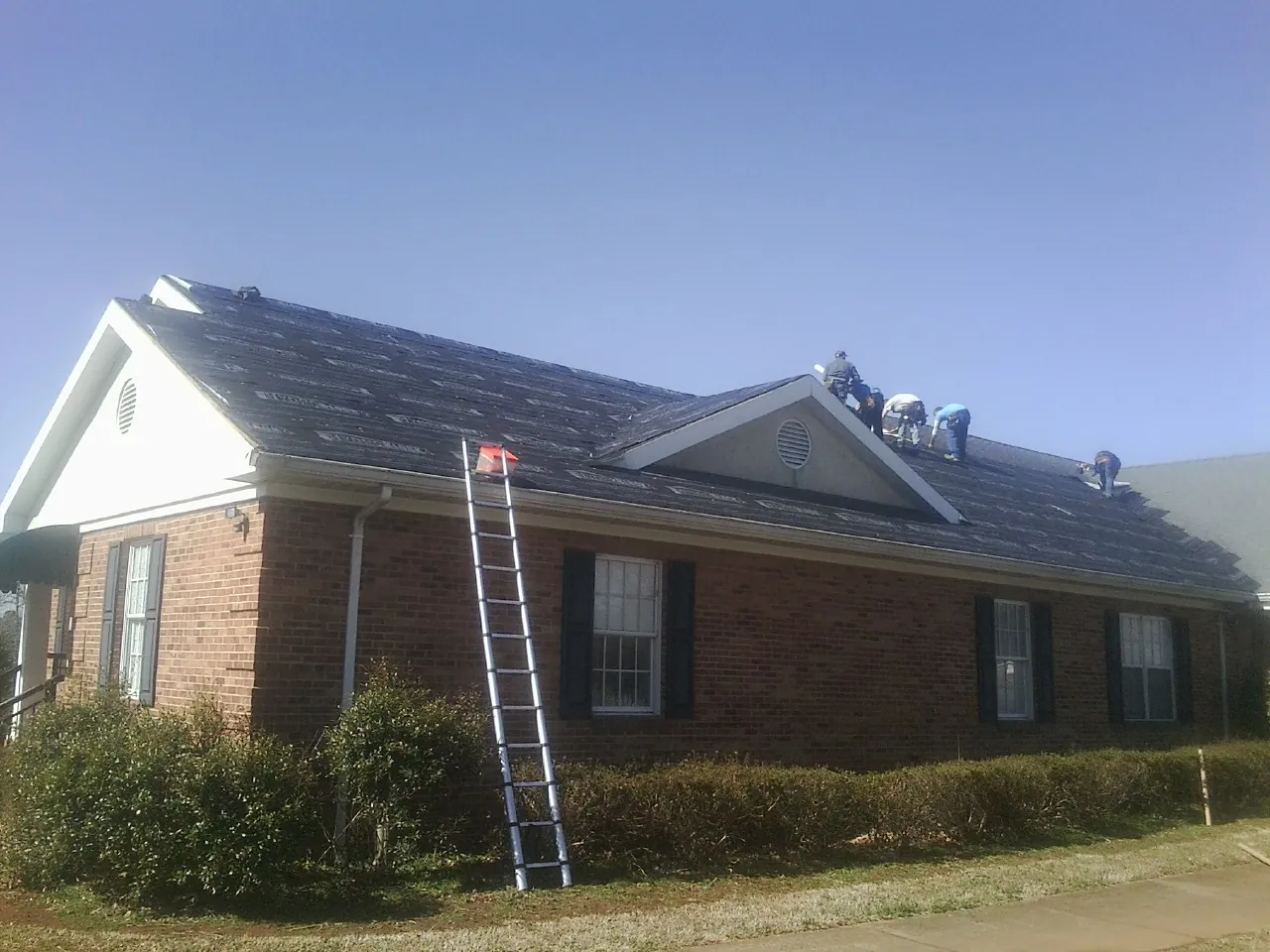 Crew tearing shingles off a brick ranch roof with a ladder set — Emergency Roof Repair work by our crew in Roper