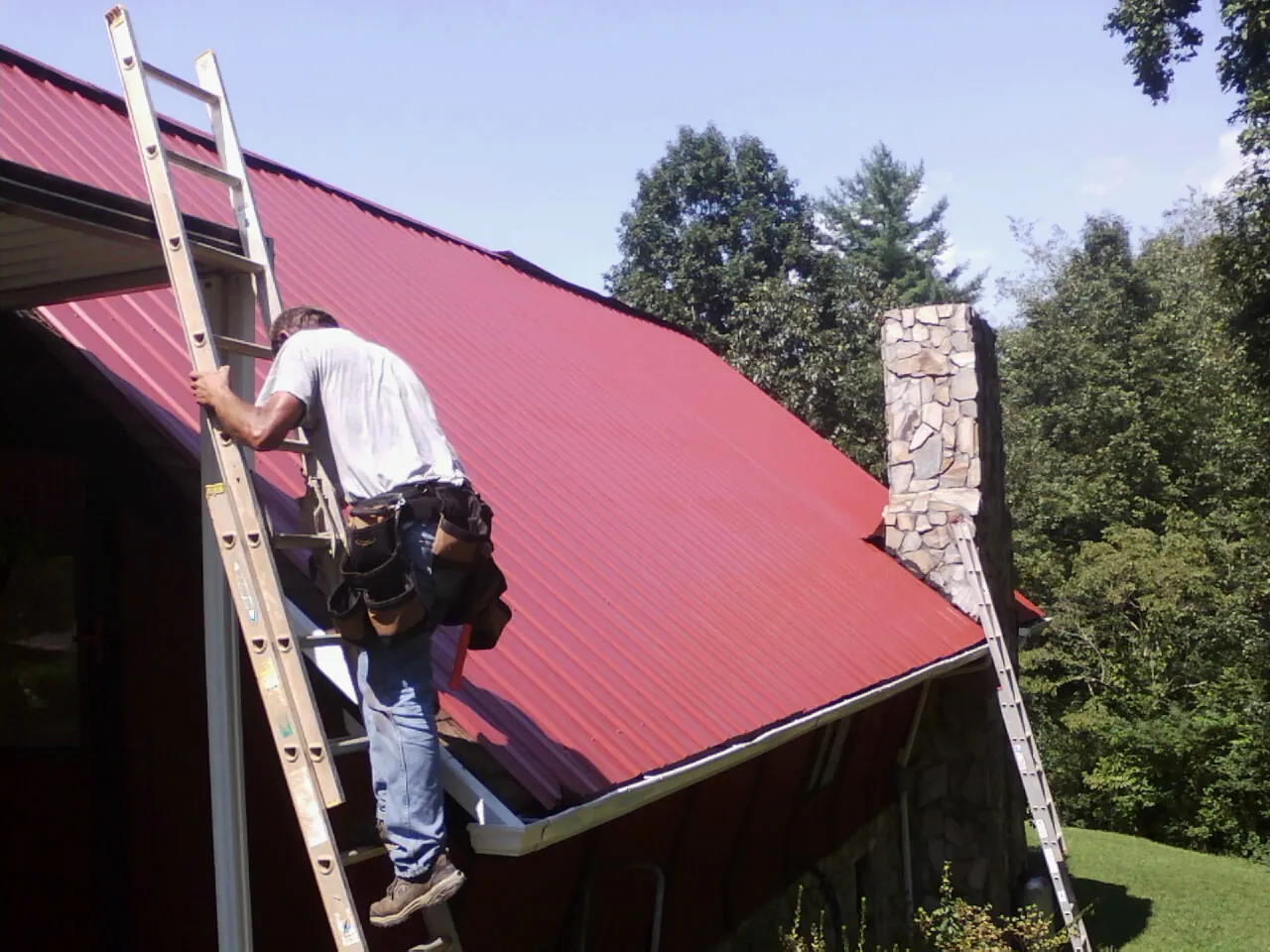 Roofer on a ladder beside a red metal roof and stone chimney — Soffit & Fascia Repair by Roofing Brothers in Roper